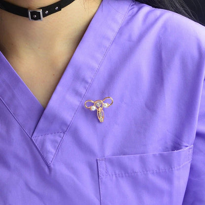 A person wears a purple scrub top with a gold uterus ovary pin—a perfect feminist gift—plus a black choker necklace featuring a silver square charm.