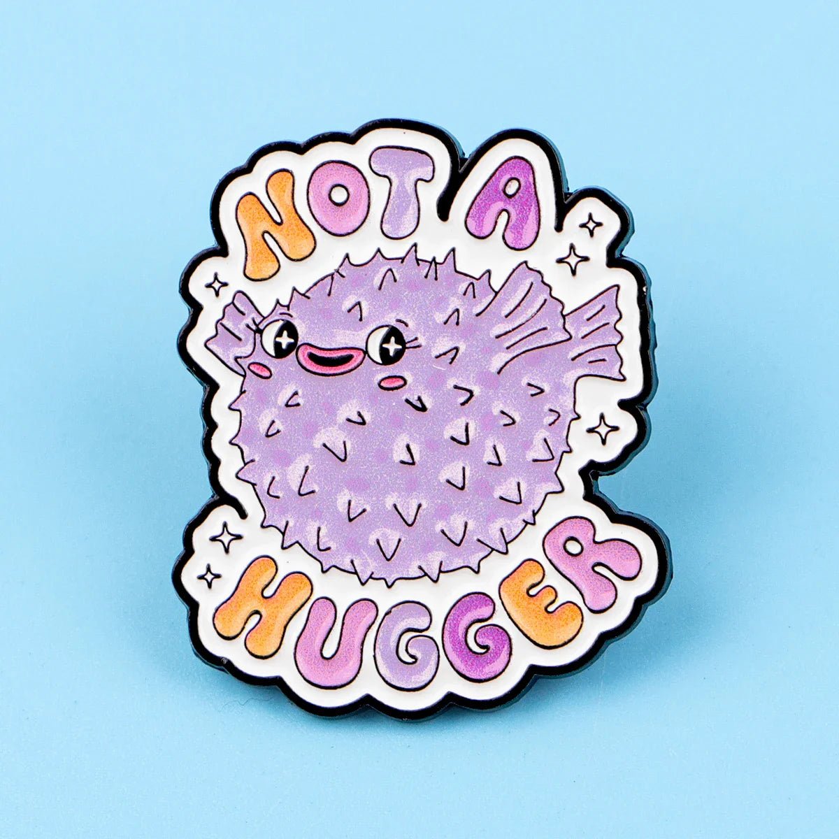 Show your pride with this Gay Skate enamel pin featuring a cute cartoon purple pufferfish and colorful Not A Hugger text on a light blue background—a playful addition to any accessories collection.