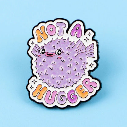 Show your pride with this Gay Skate enamel pin featuring a cute cartoon purple pufferfish and colorful Not A Hugger text on a light blue background—a playful addition to any accessories collection.