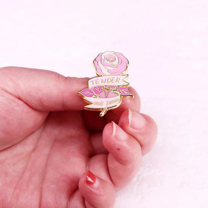 A hand displays a gold enamel pin with a pink rose and two banners that read TENDER and NOT TAME. The wearer’s painted nails complement the light pink background, making the shiny pin stand out.