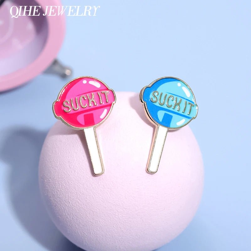 Two lollipop enamel pins, one pink and one blue, each with “SUCK IT” written on them, stand upright on a soft pink surface—add this playful Suck It pin set to your collection.