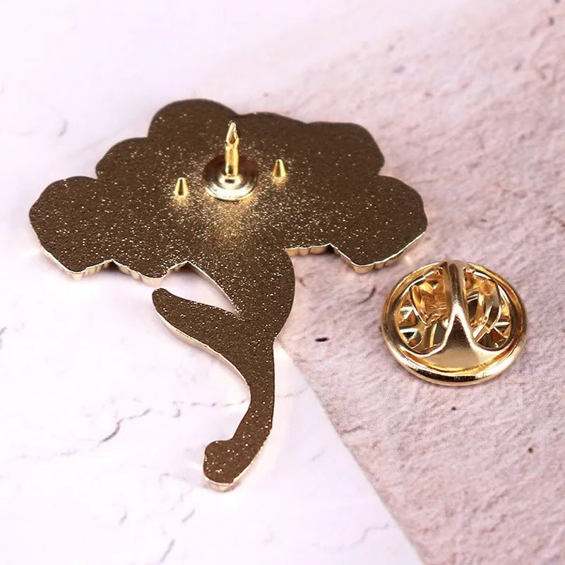 A gold-toned tree-shaped pin resembling a gothic bouquet, is shown from the back on a light textured surface with a butterfly clutch fastener nearby. The pin has a single prong and two alignment bumps.