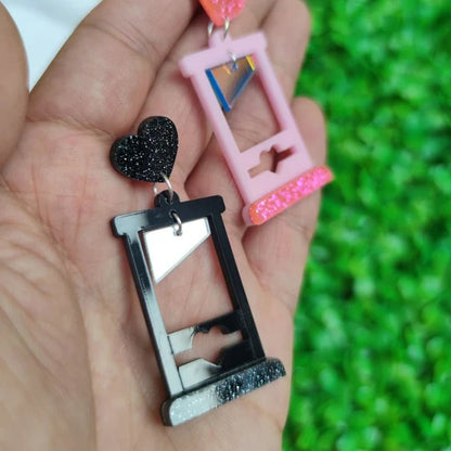 A hand holds two Gay Skate guillotine acrylic earrings—one black with a glittery heart, one pink with a matching heart—against a blurred green background.