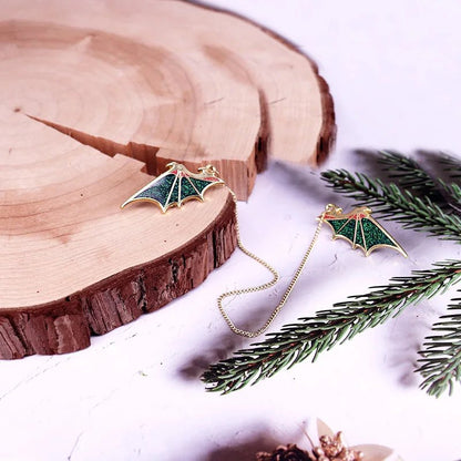 Two  bat-shaped earrings with glittery green dragon wings and gold edges rest on a wooden slab. One earring is connected by a gold chain, with pine branches nearby for decoration.