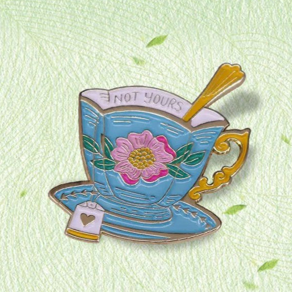 The My Cutesy Era blue floral tea cup enamel pin features a pink flower design, gold handle and spoon, matching saucer with teabag tag, NOT YOURS on the rim, and sits on a light green leaf-patterned background.