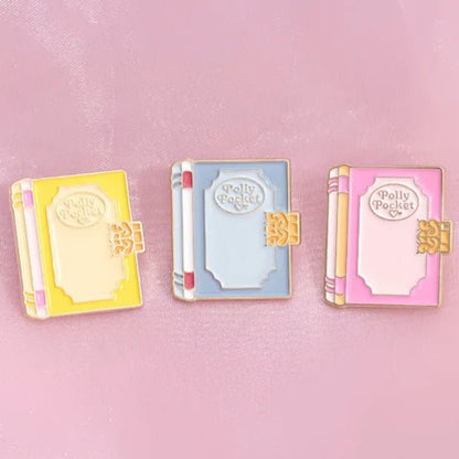 Three enamel book pins by My Cutesy Era in yellow, blue, and pink—each labeled Polly Pocket—are displayed on a light pink fabric background, perfect as charming bookworm accessories or thoughtful gifts for book lovers.