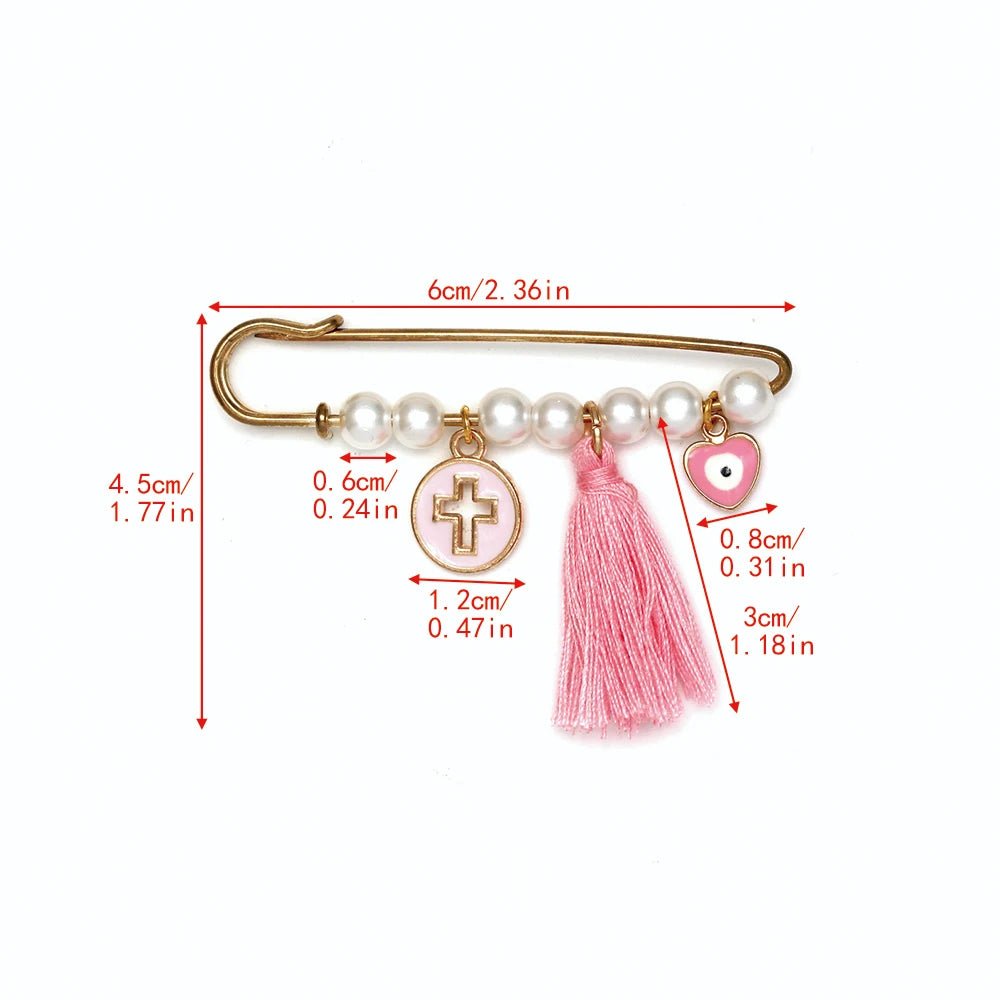 Kiss Me Laters gold safety pin brooch features five pearls, a cross charm, pink tassel, and heart charm—making it a charming baby shower gift. Red centimeter and inch lines highlight each detail against a white background.
