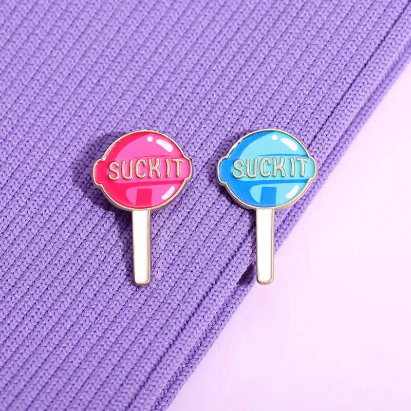 Two lollipop enamel pins in pink and blue and reading “SUCK IT,” sit on textured purple fabric. Add bold flair to your collection with a playful Suck It pin by Kiss Me Later!.