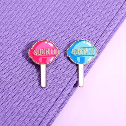 Two lollipop enamel pins in pink and blue and reading “SUCK IT,” sit on textured purple fabric. Add bold flair to your collection with a playful Suck It pin by Kiss Me Later!.