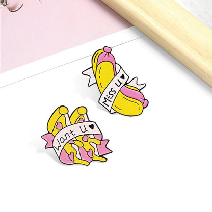 Two enamel pins shaped like peeled bananas with pink tips—one reads Miss U ♥, the other Want U ♥. These cute yellow and pink pins rest on a white surface near a light wooden object.