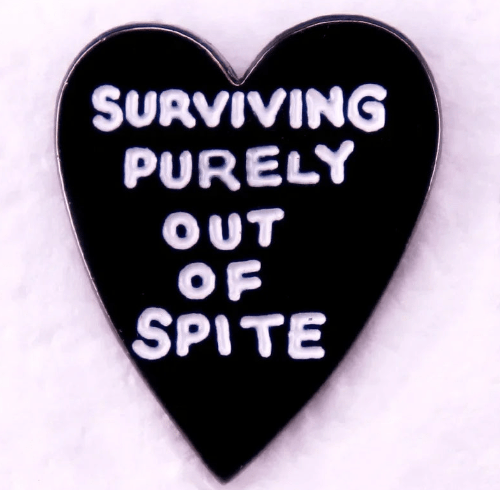 A black heart-shaped pin with white SURVIVING PURELY OUT OF SPITE text sits next to a red love heart enamel pin on a light pink background.