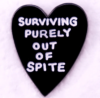 A black heart-shaped pin with SURVIVING PURELY OUT OF SPITE in white text rests on a light pink background—an ideal match for your favorite red love heart enamel pin.