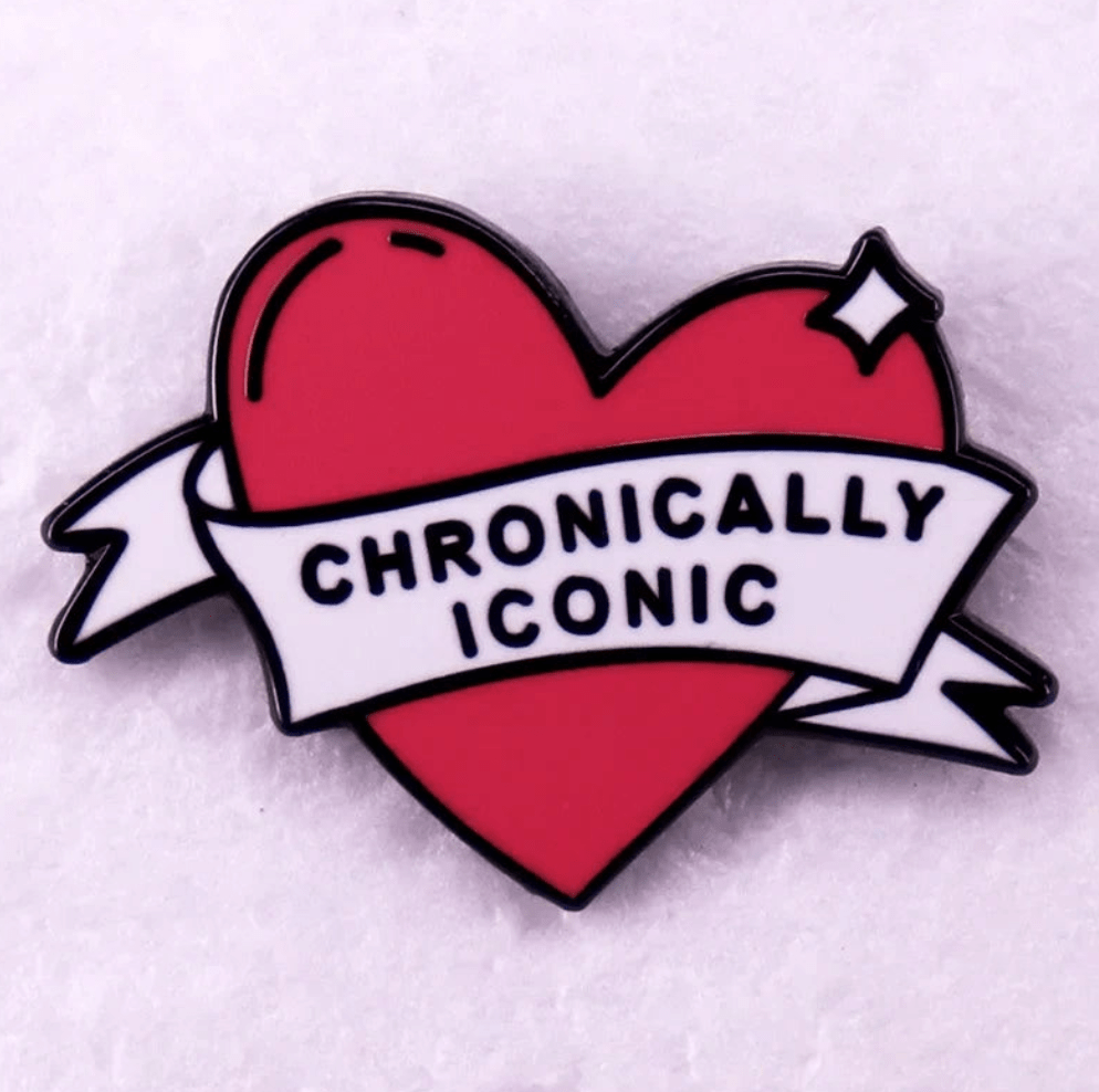 RED love heart enamel pin with a white banner that says “CHRONICALLY ICONIC” in black letters, shown on a soft, light background.