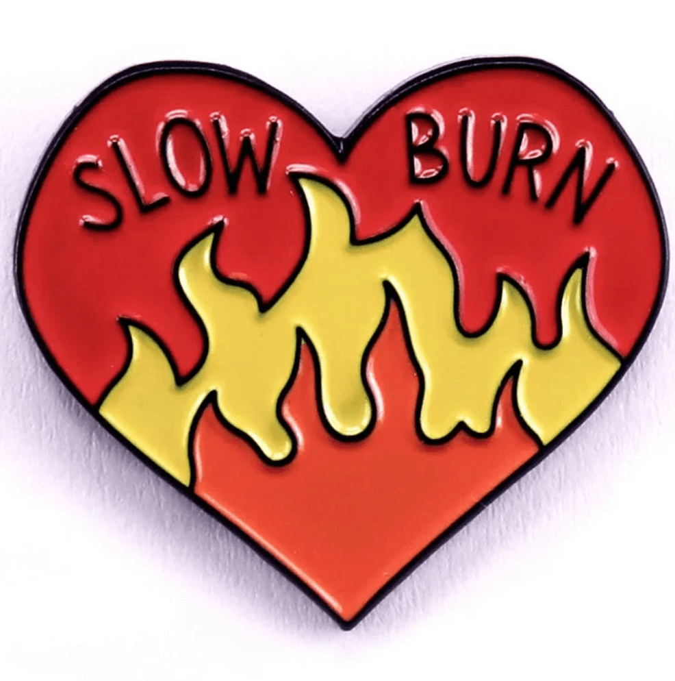 Red heart enamel pin with SLOW BURN at the top, yellow flames rising from the bottom, and a vibrant red-orange background.