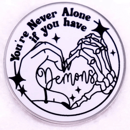 Round white enamel pin features a human and skeleton hand forming a heart around Demons, surrounded by stars, with the phrase You’re Never Alone if you have Demons.