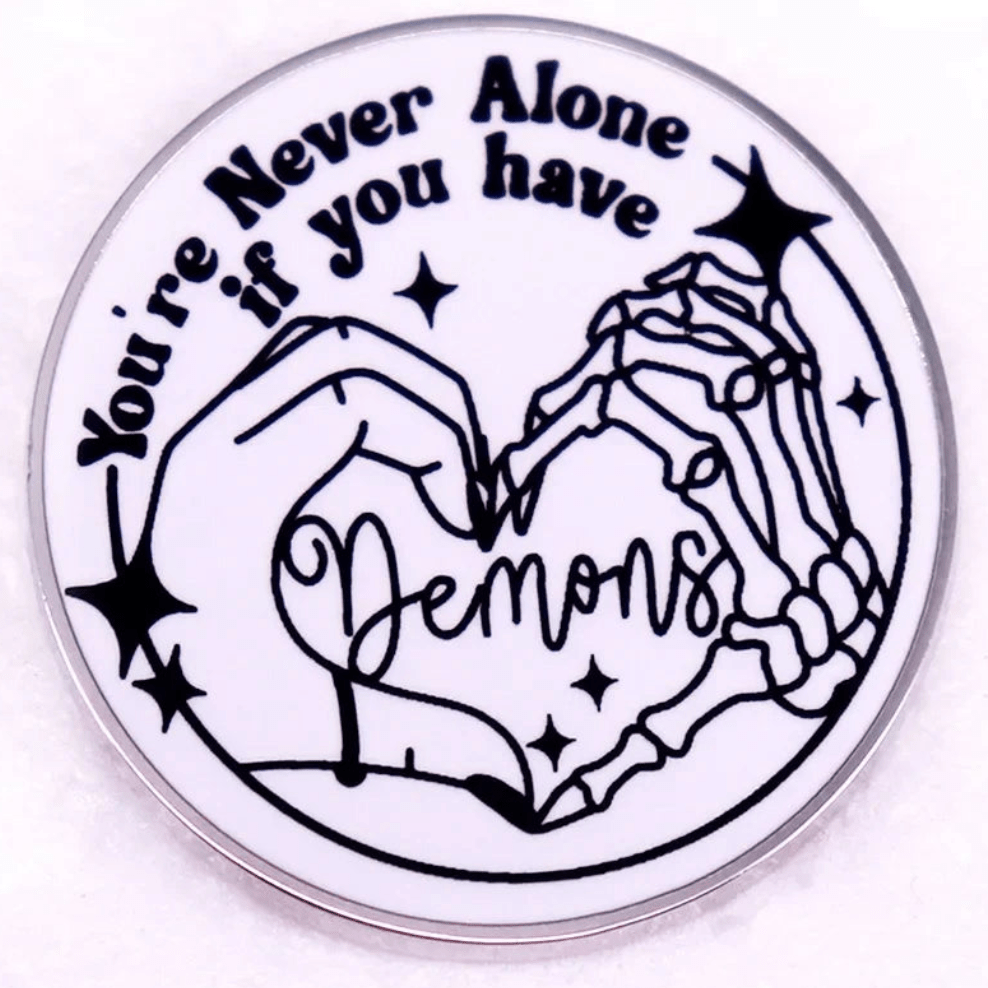 Enamel pin is a circular red heart with You’re Never Alone if you have Demons, showing a human and skeleton hand forming a heart, plus stars and sparkles.