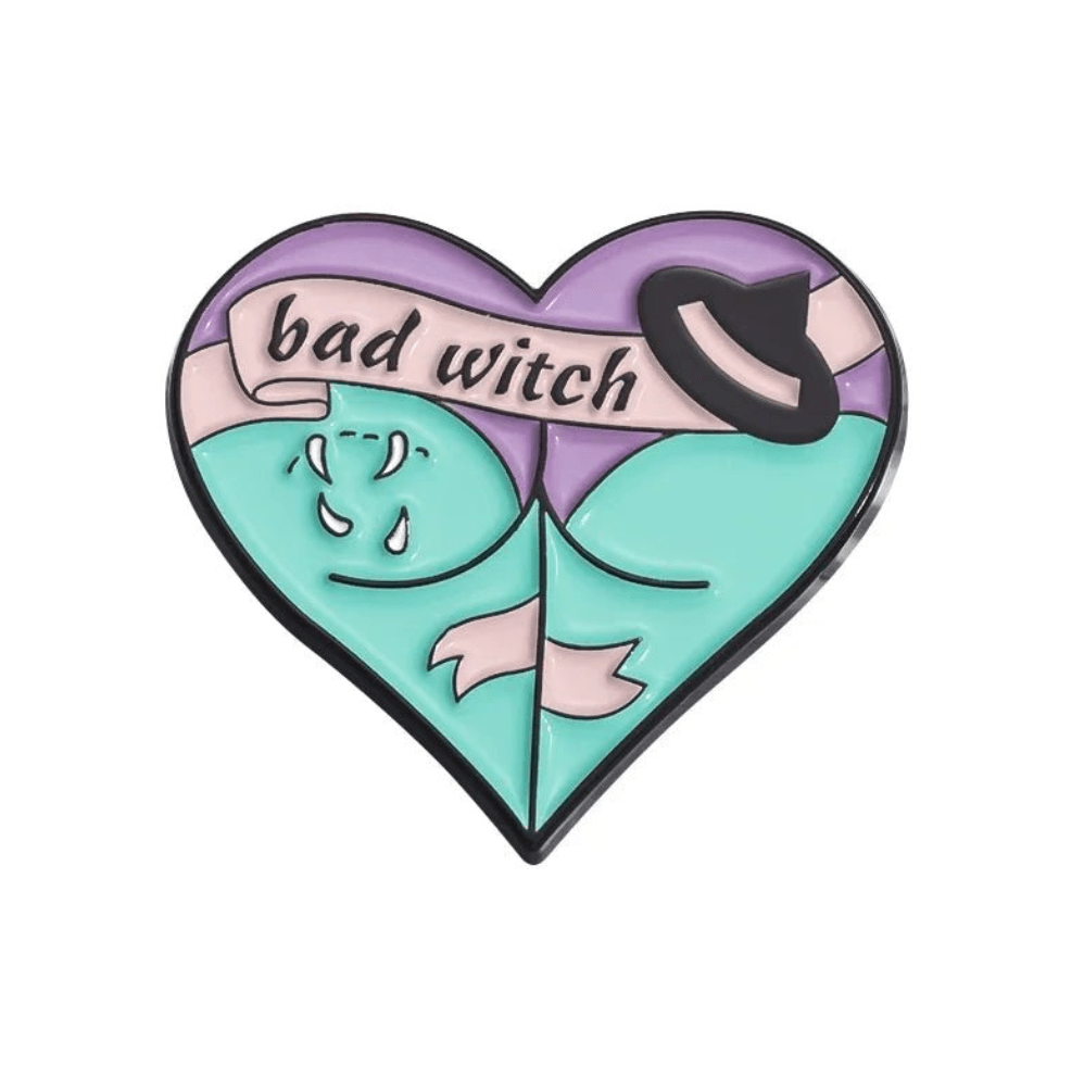 Heart-shaped enamel pin in pastel colors, with bad witch on a banner, a witch hat, gothic details, and a stylized woman’s chest and cleavage.