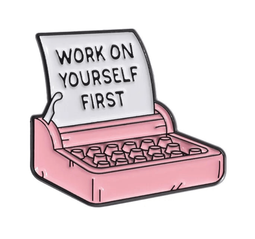 A pink typewriter sits next to a tiny espresso machine, with a sheet of paper emerging that reads, WORK ON YOURSELF FIRST.
