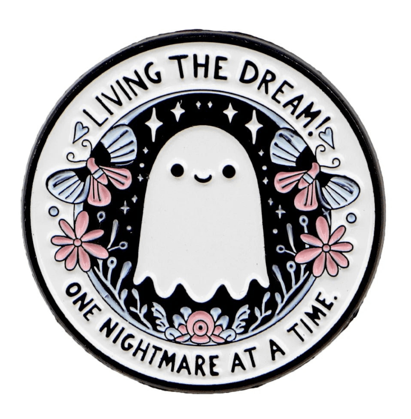 Enamel pin that features a cute ghost surrounded by flowers and moths, with the text Living The Dream One Nightmare At A Time. This whimsical enamel pin has pink accents on a black background, ideal for collectors.