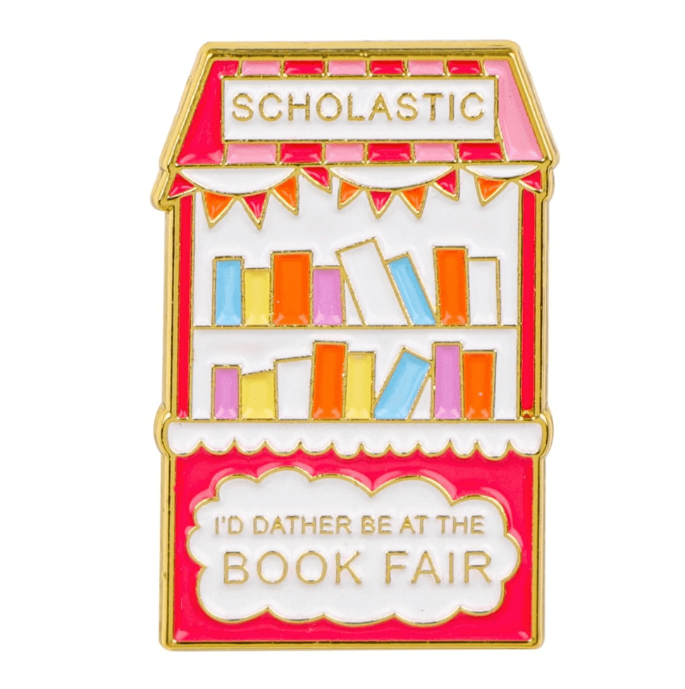 A vibrant enamel pin shaped like a book fair stand, featuring SCHOLASTIC on top, colorful books, and ID RATHER BE AT THE BOOK FAIR—a must-have for enamel pin collectors and book lovers.