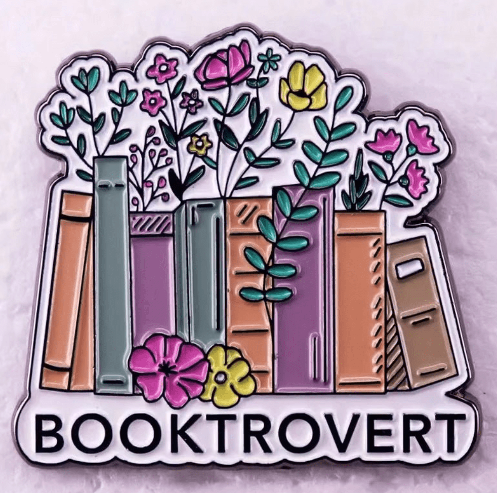 A colorful enamel pin by featuring books with flowers and leaves, above BOOKTROVERT in bold—a perfect badge for any romance reader and book lover.