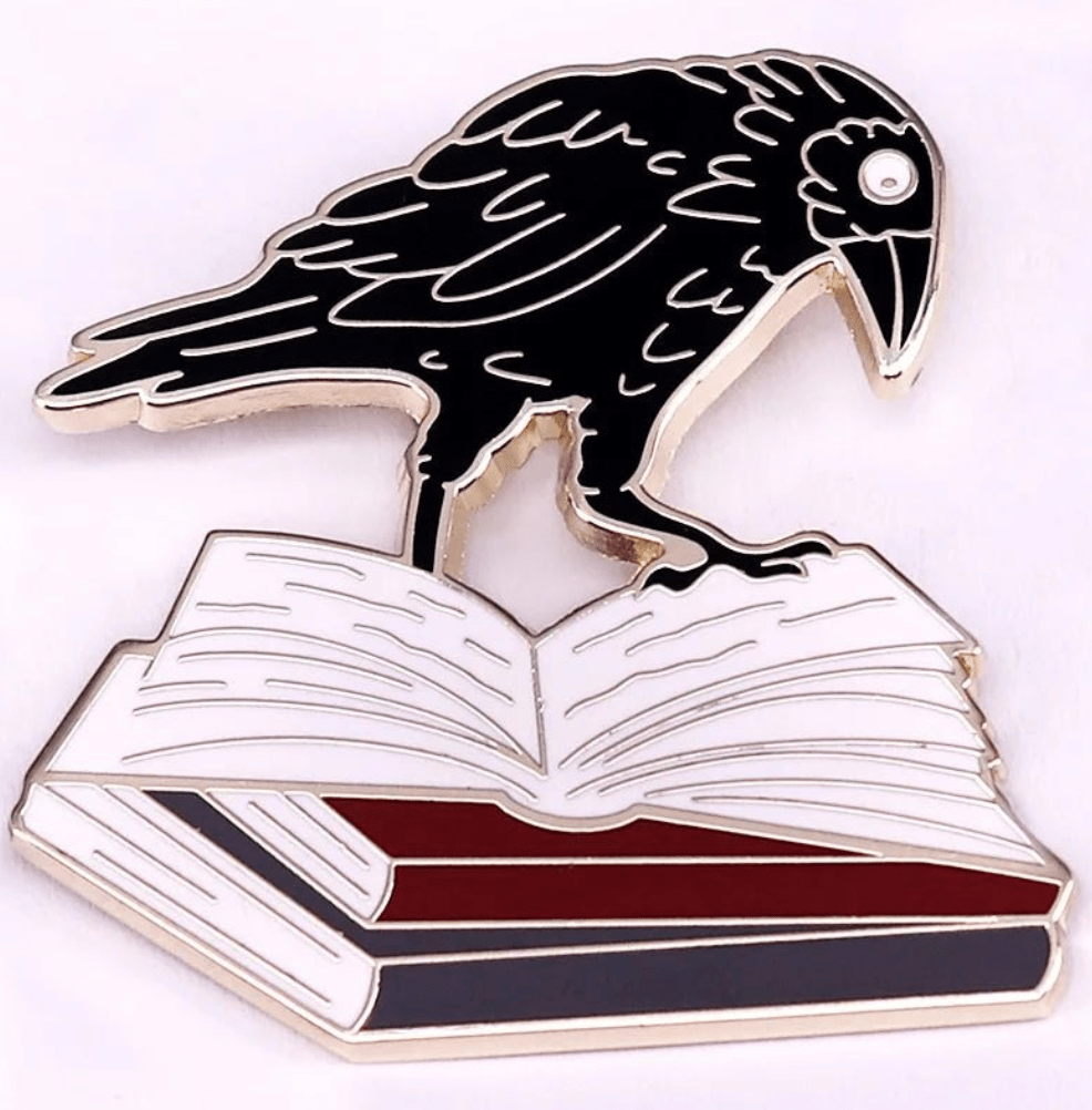 A black raven stands atop an open book with red, white, and black pages on this striking enamel pin—a perfect accessory for romance readers and book lover badge collections.