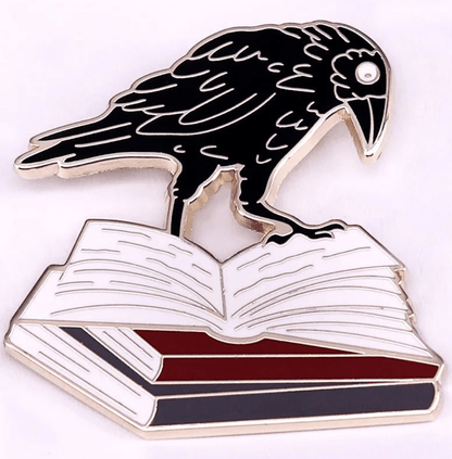 A black raven stands atop an open book with red, white, and black pages on this striking enamel pin—a perfect accessory for romance readers and book lover badge collections.