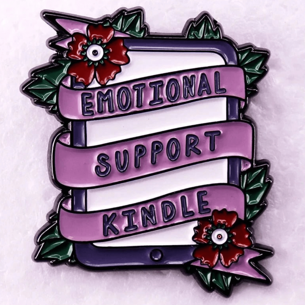 Enamel pin features a Kindle wrapped in a pink ribbon with red flowers and green leaves. The ribbon reads “EMOTIONAL SUPPORT KINDLE” in bold black letters—perfect for romance reader book lover badges.