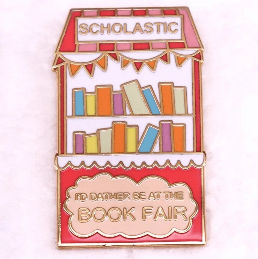 A colorful enamel pin shaped like a book fair booth with books on shelves. SCHOLASTIC tops it, and ID RATHER BE AT THE BOOK FAIR is in gold—ideal for book lovers or romance readers’ badges.