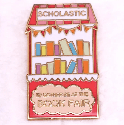 A colorful enamel pin shaped like a book fair booth with books on shelves. SCHOLASTIC tops it, and ID RATHER BE AT THE BOOK FAIR is in gold—ideal for book lovers or romance readers’ badges.