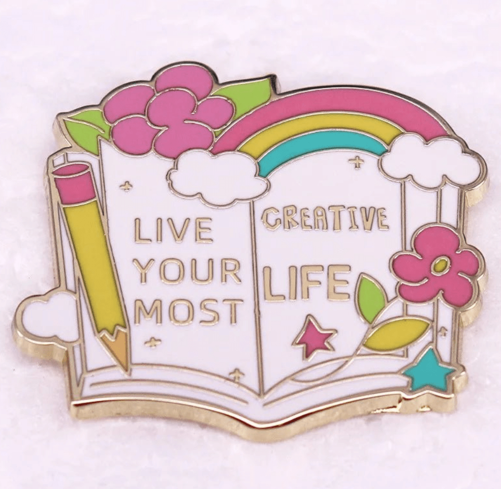 Colorful enamel pin features an open book with flowers, pencil, clouds, and rainbow, plus Live your most creative life in gold—an ideal badge for every Romance Reader.