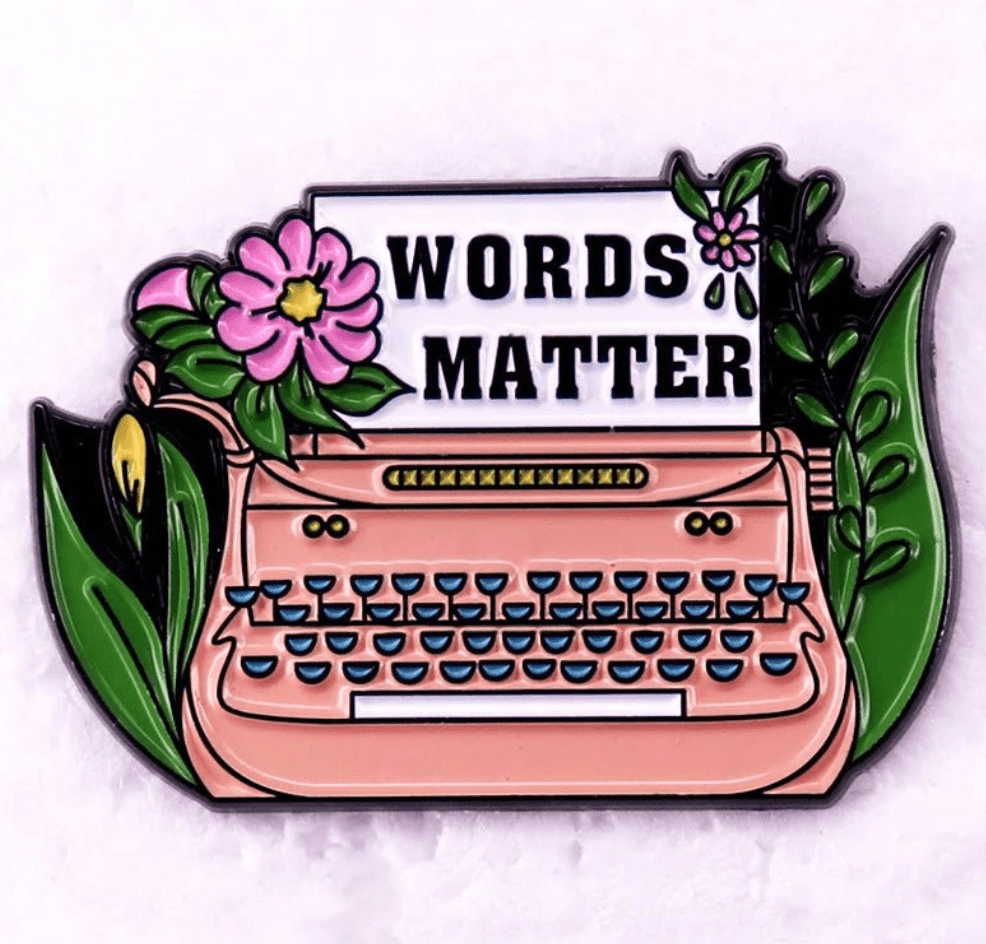 A Kiss Me Later pin featuring a pink typewriter with green keys and flowers, holding a “WORDS MATTER” sheet. Ideal for Romance Reader enamel pin fans, this vibrant design stands out on a pale background.