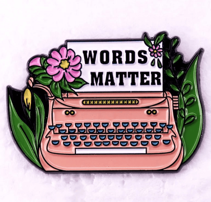 A Kiss Me Later pin featuring a pink typewriter with green keys and flowers, holding a “WORDS MATTER” sheet. Ideal for Romance Reader enamel pin fans, this vibrant design stands out on a pale background.