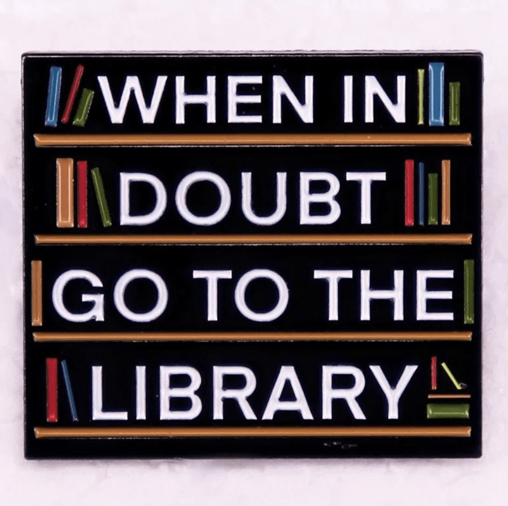 A Kiss Me Later sign features colorful books on shelves with When in doubt go to the library in bold white letters on a black background—perfect inspiration for enamel pins or book lover badge designs.