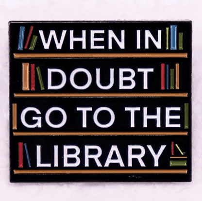 A Kiss Me Later sign features colorful books on shelves with When in doubt go to the library in bold white letters on a black background—perfect inspiration for enamel pins or book lover badge designs.
