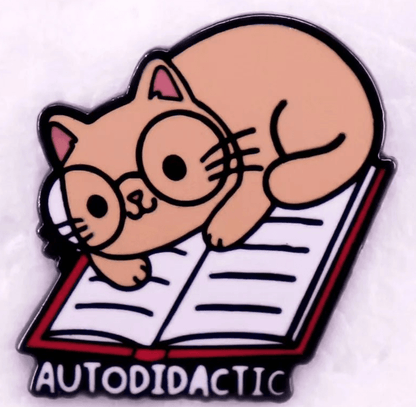 A cartoon cat with large glasses lounges on an open book above the bold word “AUTODIDACTIC.” This quirky enamel pin is perfect for book lovers or anyone embracing lifelong learning.