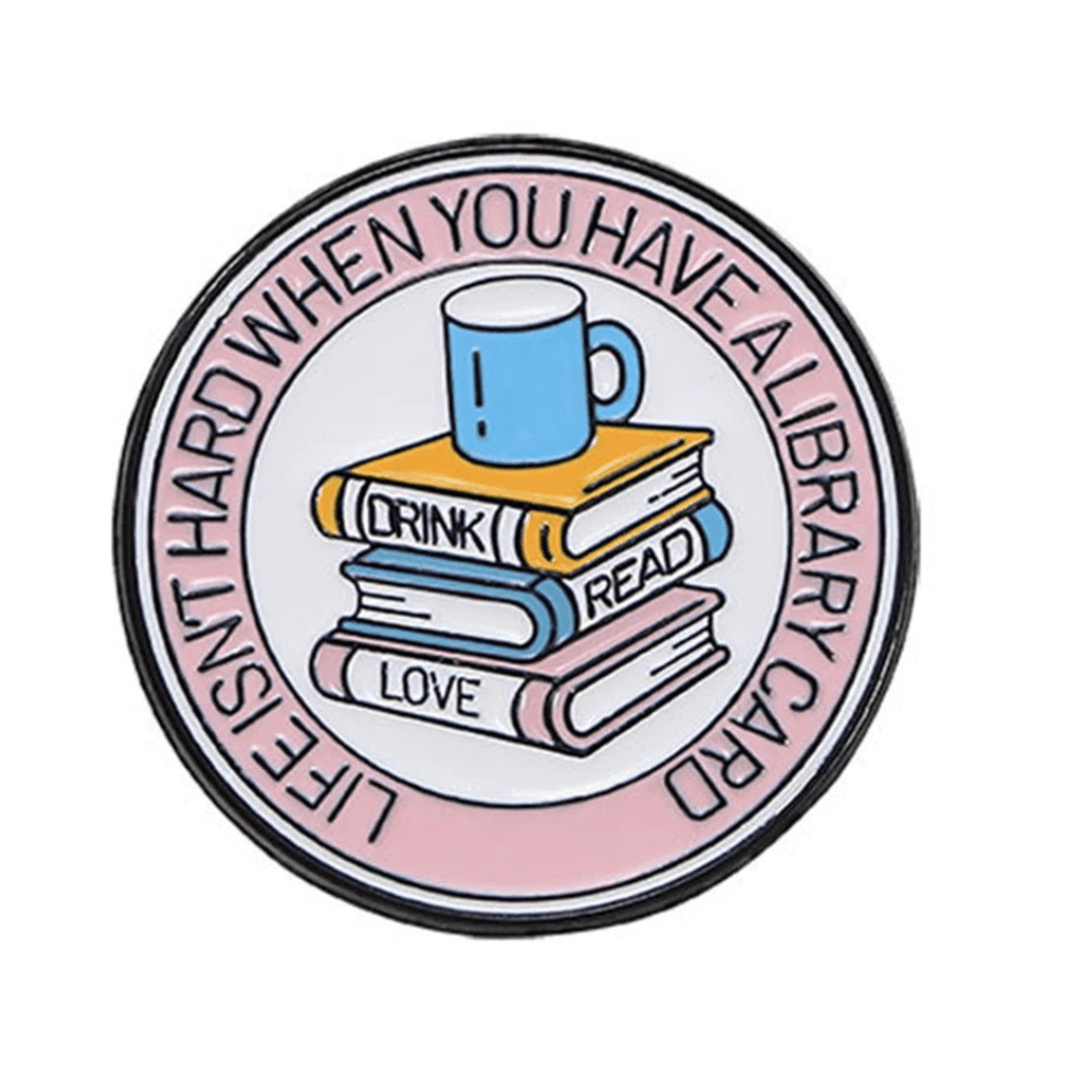 Round book lover badge features a blue mug on three books labeled DRINK, READ, LOVE. Rim text reads LIFE ISN’T HARD WHEN YOU HAVE A LIBRARY CARD”—a must-have for Romance Readers or enamel pin collectors.
