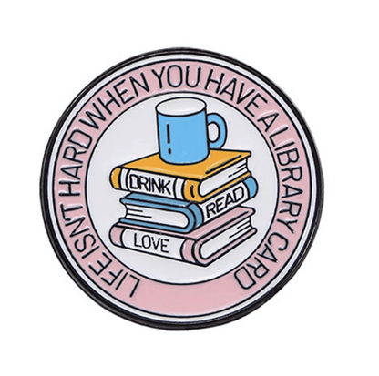 Round book lover badge features a blue mug on three books labeled DRINK, READ, LOVE. Rim text reads LIFE ISN’T HARD WHEN YOU HAVE A LIBRARY CARD”—a must-have for Romance Readers or enamel pin collectors.