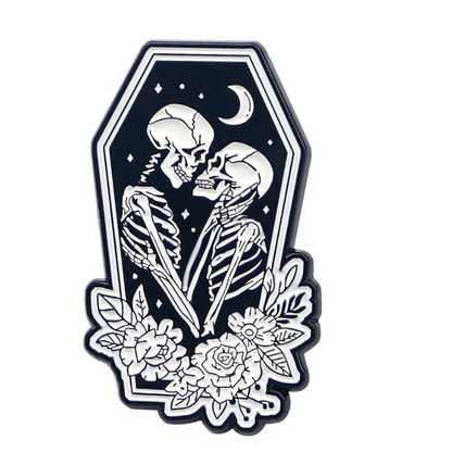 bold black and white Skeleton Lovers enamel pin shows two skeletons embracing in a coffin-shaped frame, with a crescent moon, stars, and floral accents—capturing striking outlines and timeless romance.