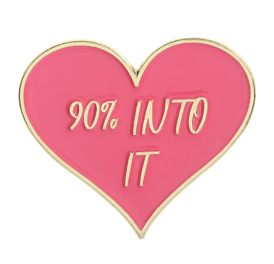 Enamel pin is pink, heart-shaped with gold edges, featuring red and gold details and 90% INTO IT in gold letters.