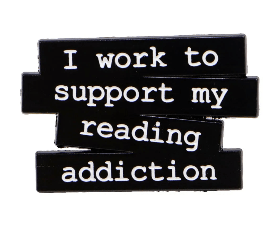 black pin features white text, I work to support my reading addiction, on staggered blocks, accented with a small red heart enamel detail.