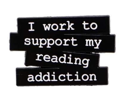 black pin features white text, I work to support my reading addiction, on staggered blocks, accented with a small red heart enamel detail.