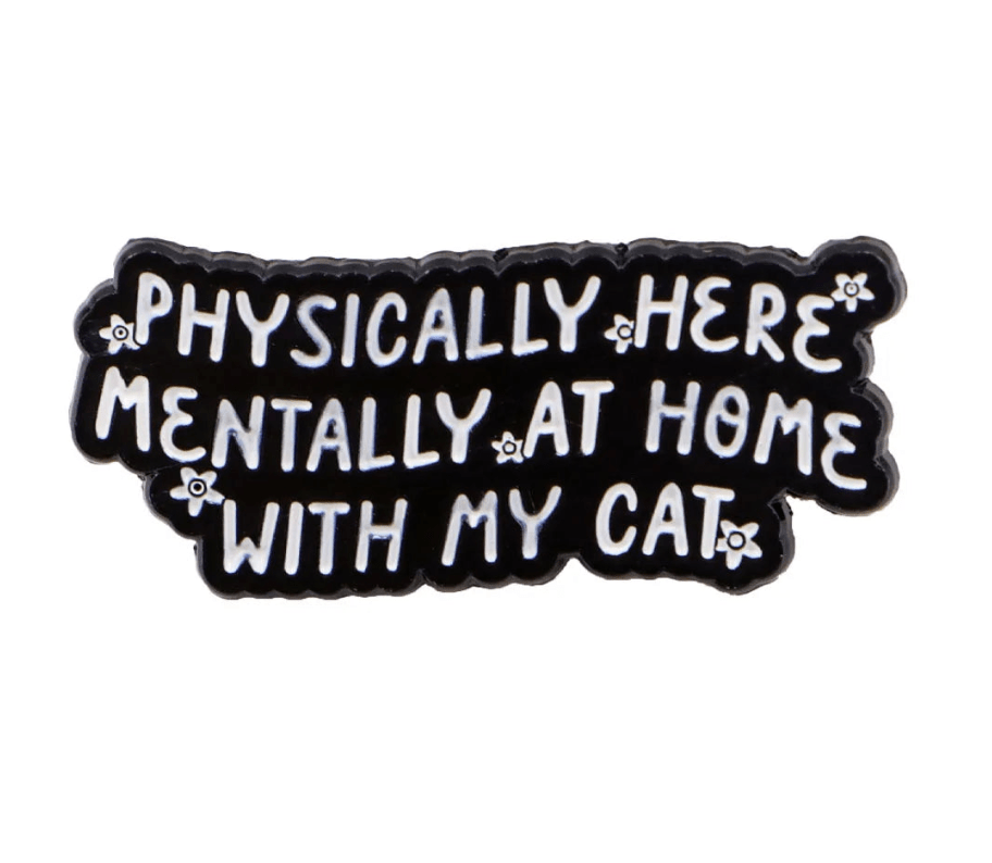 A black pin with white text reads, Physically here, mentally at home with my cat. It features a red love heart enamel pin and small white flowers along the edges.