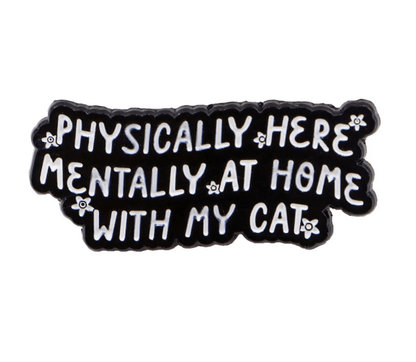 A black pin with white text reads, Physically here, mentally at home with my cat. It features a red love heart enamel pin and small white flowers along the edges.