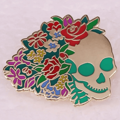 Enamel pin shaped like a skull, decorated with bright red roses, purple flowers, and yellow leaves arranged on one side against a light background.