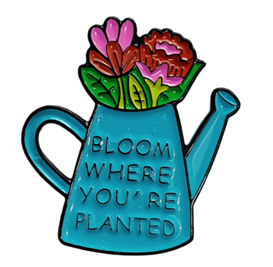 A charming black and gold enamel pin by My Cutesy Era features a blue watering can with colorful flowers and the phrase BLOOM WHERE YOU’RE PLANTED—perfect for Black Sheep Club members.