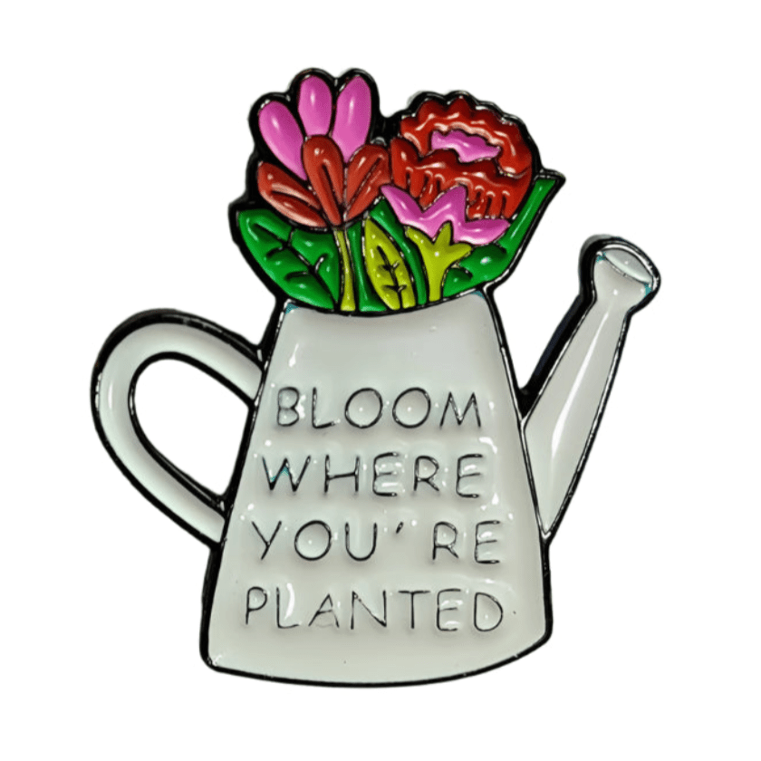 A white enamel pin by My Cutesy Era, shaped like a watering can with colorful flowers and the phrase BLOOM WHERE YOU’RE PLANTED on the front.