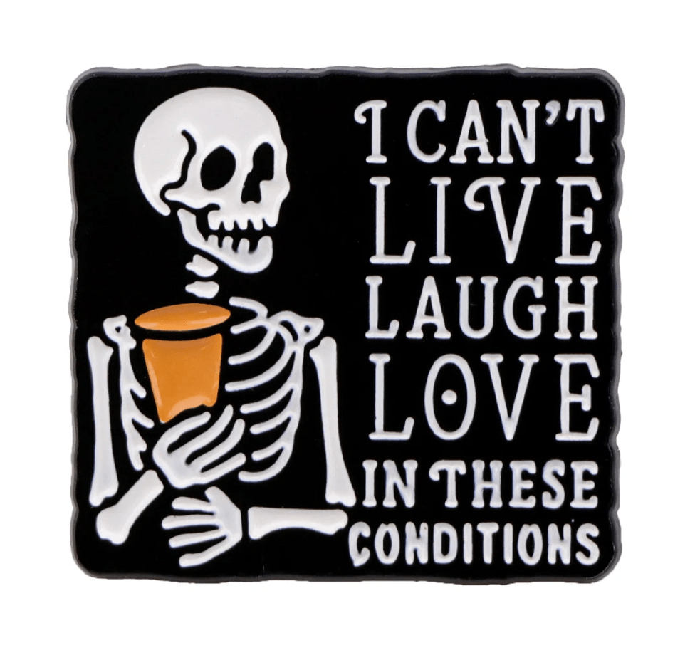 A cartoon skeleton with a drink stands by the text I CANT LIVE LAUGH LOVE IN THESE CONDITIONS on black—perfect for fans of gothic pins. From Kiss Me Later.