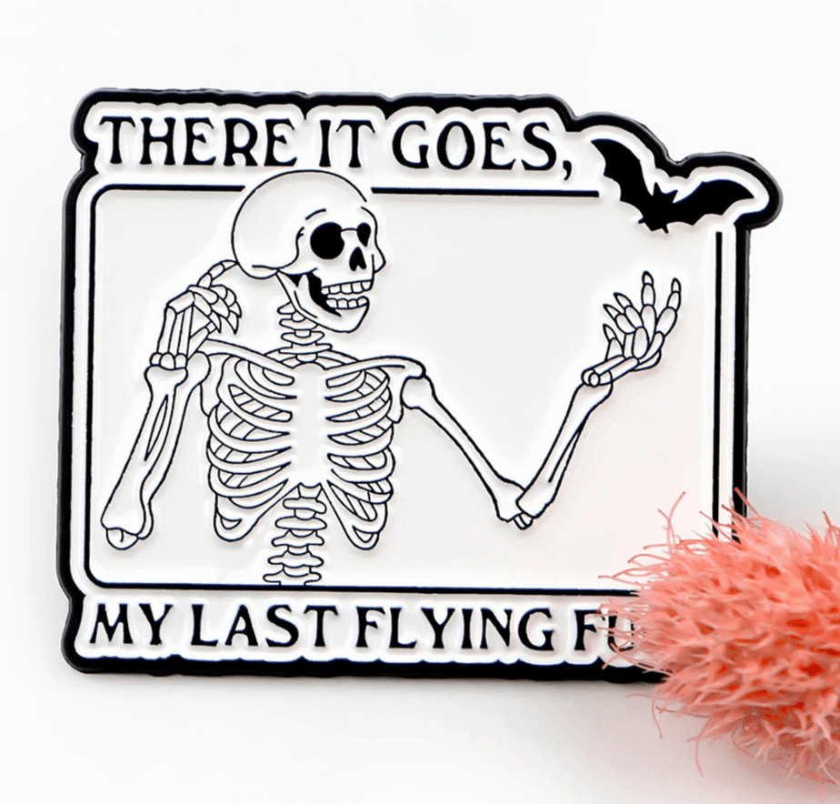 A skeleton with one arm raised and a bat overhead is framed by “There it goes, my last flying fu.” A fuzzy pink object sits in the lower right. Perfect for skeleton pin fans. Brand: Kiss Me Later.