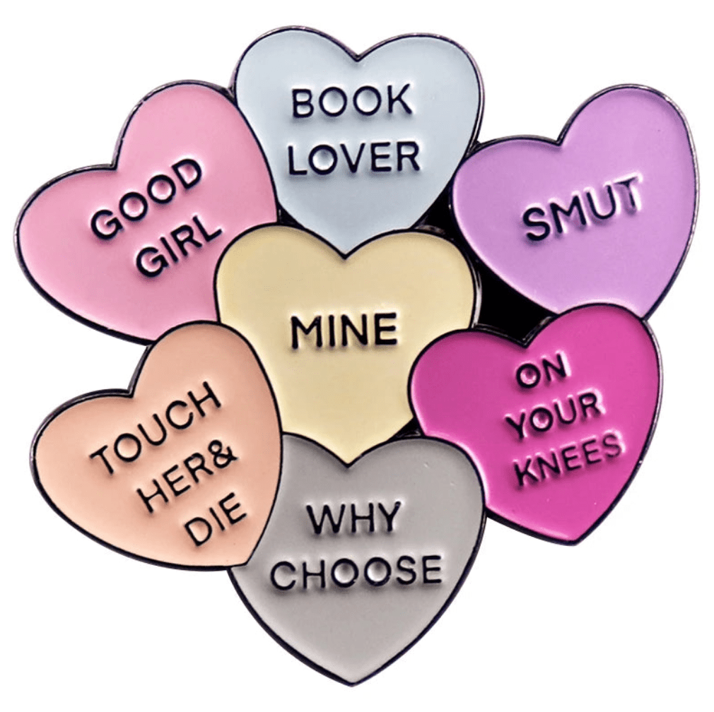 A cluster of seven pastel love hearts enamel pins, each with unique text: GOOD GIRL, SMUT pin, BOOK LOVER, ON YOUR KNEES, WHY CHOOSE, TOUCH HER & DIE, and MINE at the center.